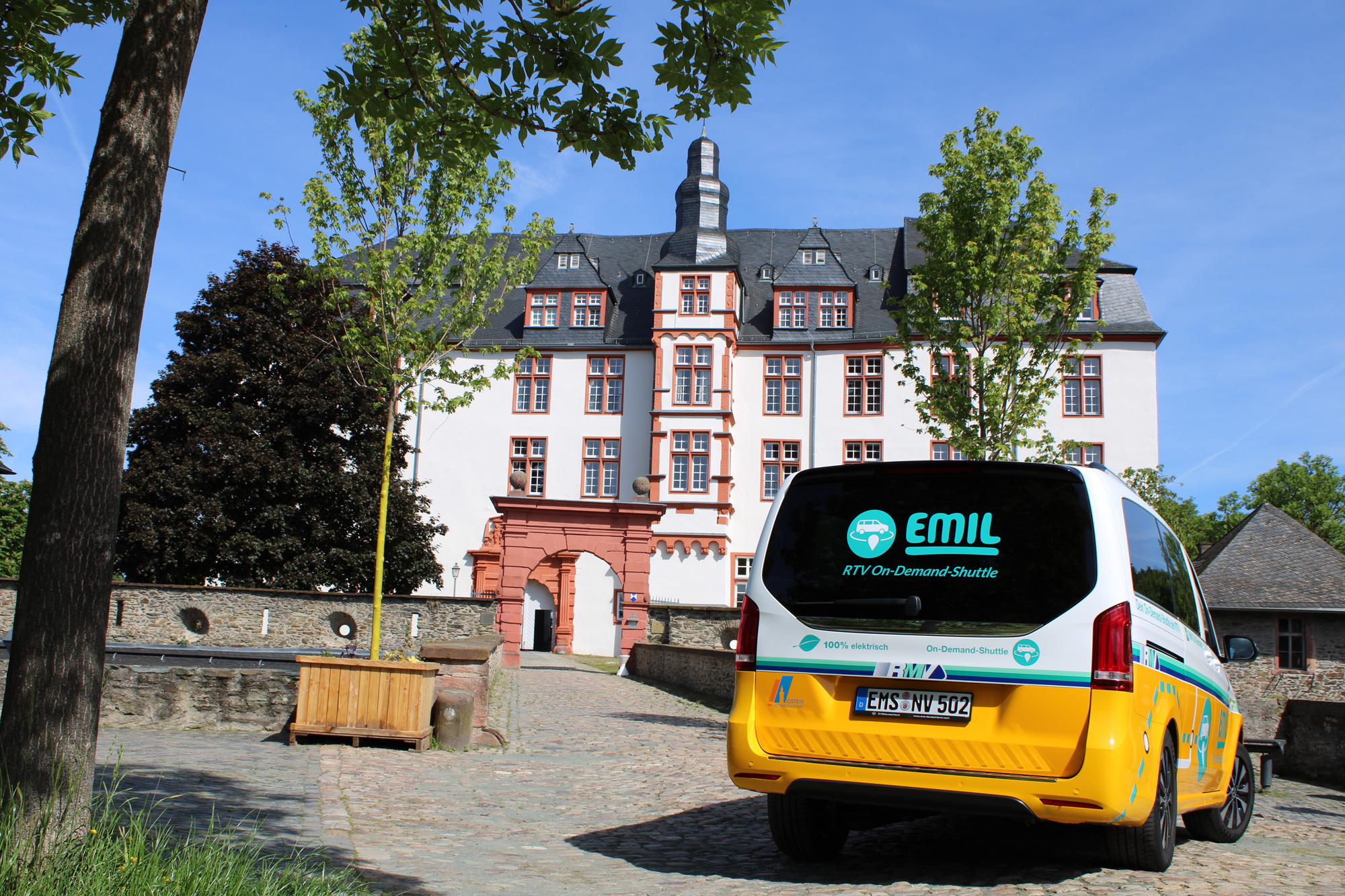 Das elektrisch betriebene On-Demand-Shuttle „EMIL“ der Rheingau-Taunus-Verkehrsgesellschaft steht vor dem historischen Idsteiner Schloss mit weiß-roter Fassade und markantem Schieferdach.