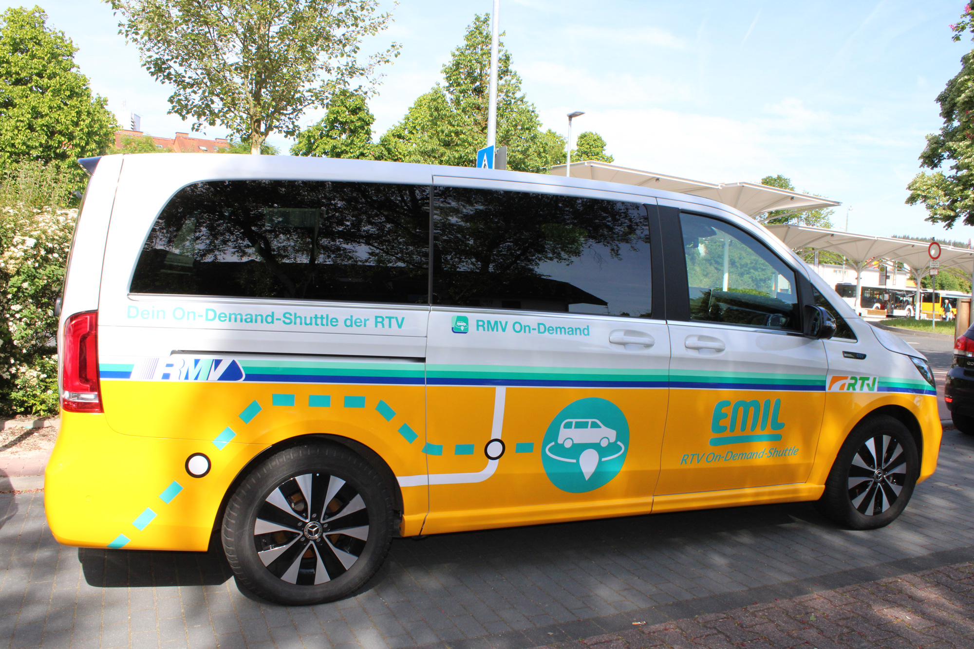 Ein gelb-weißes EMIL-On-Demand-Shuttle der Rheingau-Taunus-Verkehrsgesellschaft steht am Straßenrand vor dem überdachten Busbahnhof. Auf der Fahrzeugseite sind RMV- und RTV-Logos sowie eine stilisierte Routenlinie sichtbar.